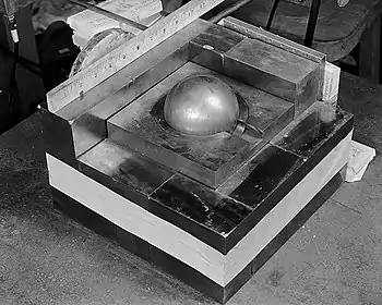 A square base of metal blocks, with a smaller square of metal on the top in the center, a metal ball (the "core") contained in its center. A ruler along one side of the base shows it is roughly 10.5 inches (270&nbsp;mm) square.